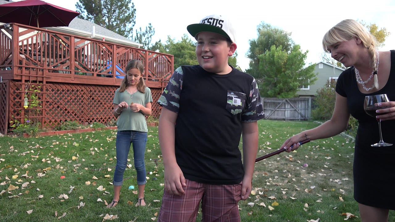 Boy with croquet mallet in a backyard while a girl and woman stand nearby.