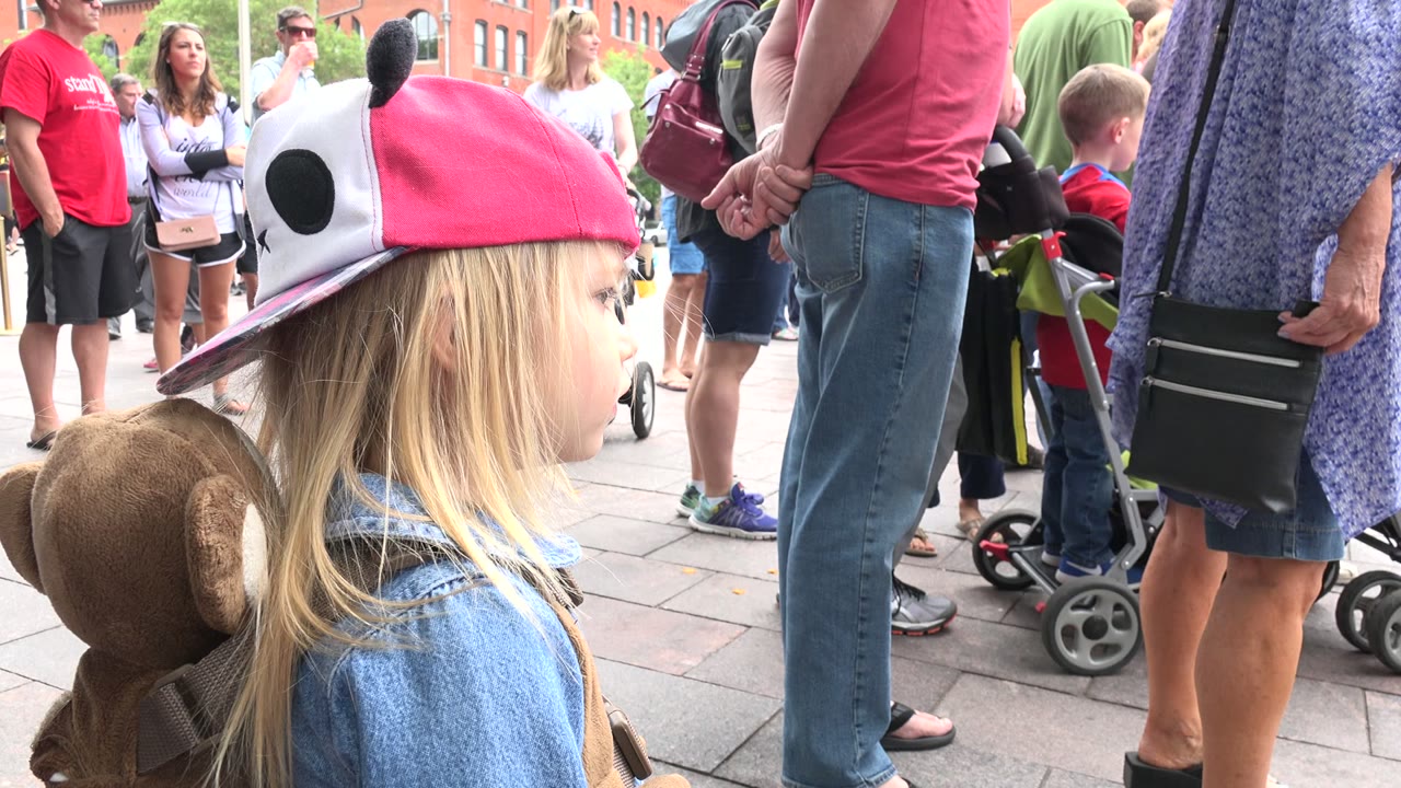 A young blonde child in a pink panda cap and denim shirt stands in side profile in a Grand Junction, Colorado plaza, with adults and strollers behind her.