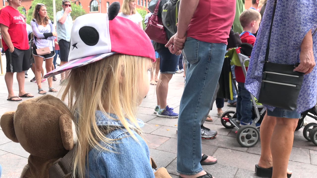 The child remains in profile at close range in Grand Junction, Colorado, with the plush monkey backpack visible and a line of adults and strollers in the background.