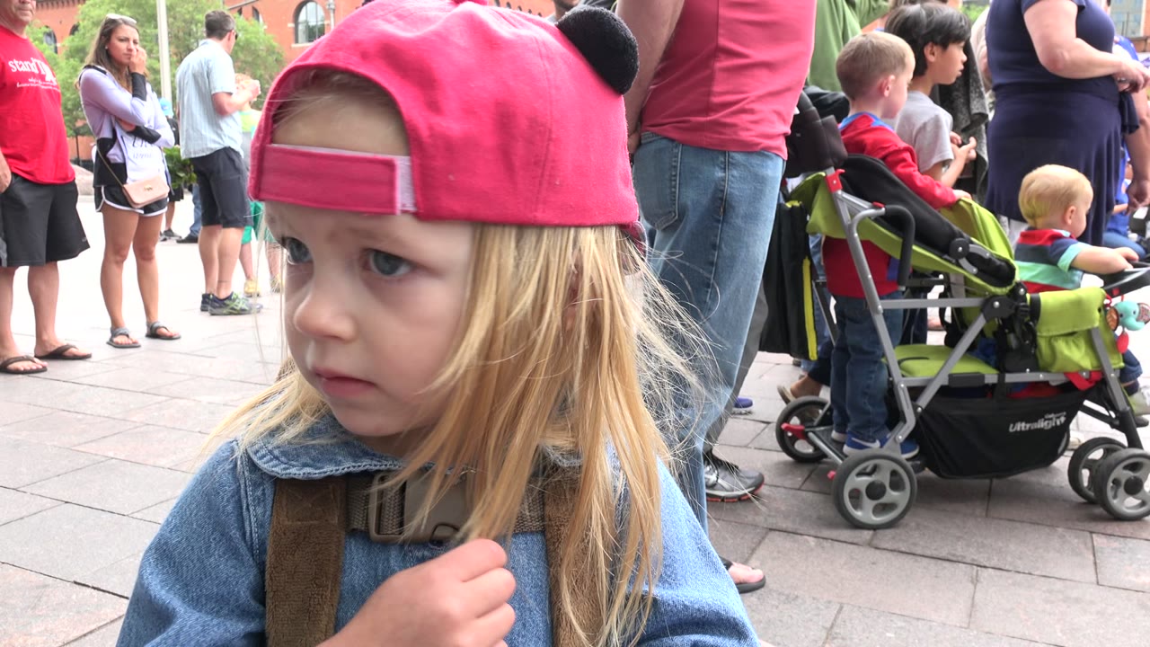 Young child in a backward pink cap looks left in a crowded outdoor plaza.