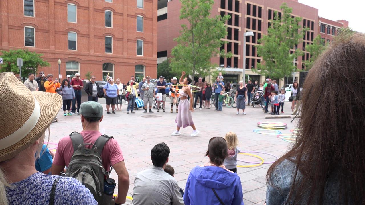 In Grand Junction, Colorado, the performer extends her arms near several hoops as spectators line the plaza, with foreground audience members partly blocking the lower frame.