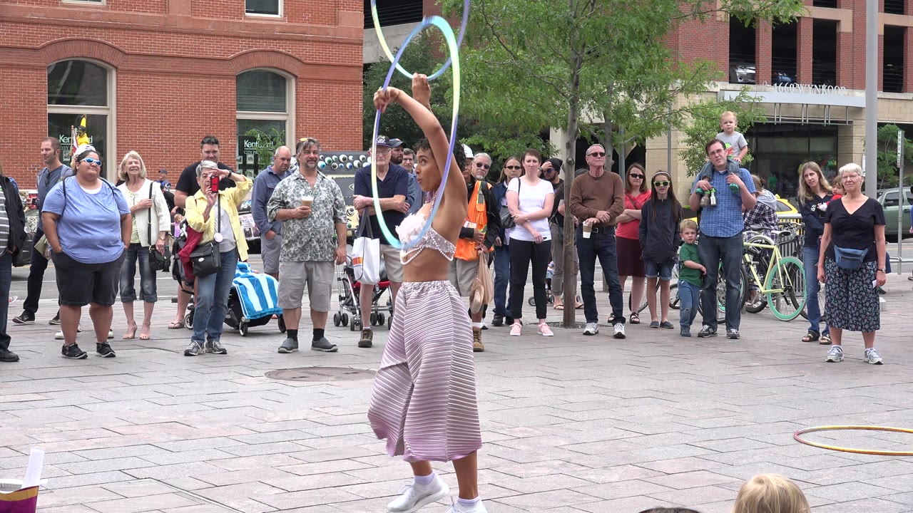The Grand Junction street performer holds two hoops nearly vertical beside her body, with spectators lined up behind her near a brick building.