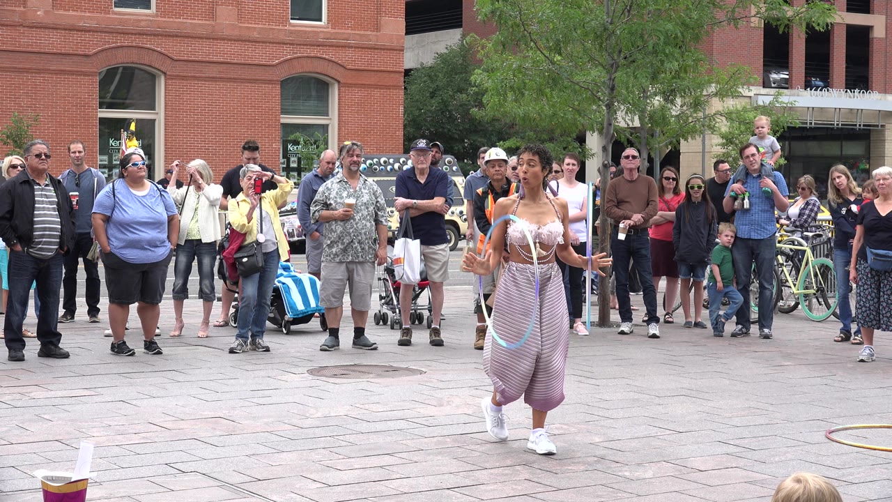 At the start of the clip in Grand Junction, Colorado, a woman performer faces forward with hoops near her hands while spectators stand behind her on a paved plaza.