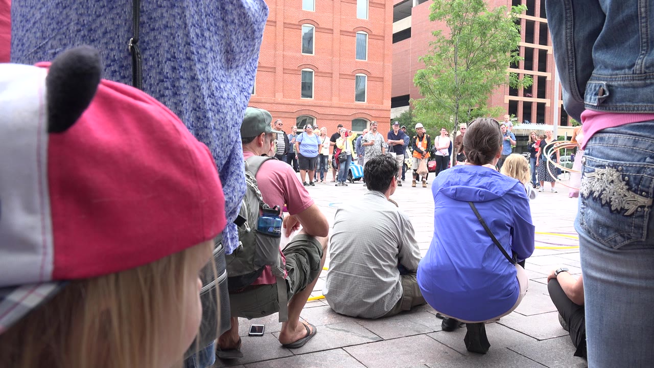 Grand Junction, Colorado crowd watches a street performance from behind seated spectators, with a child in a pink-and-white cap close to camera and red brick buildings behind the circle.