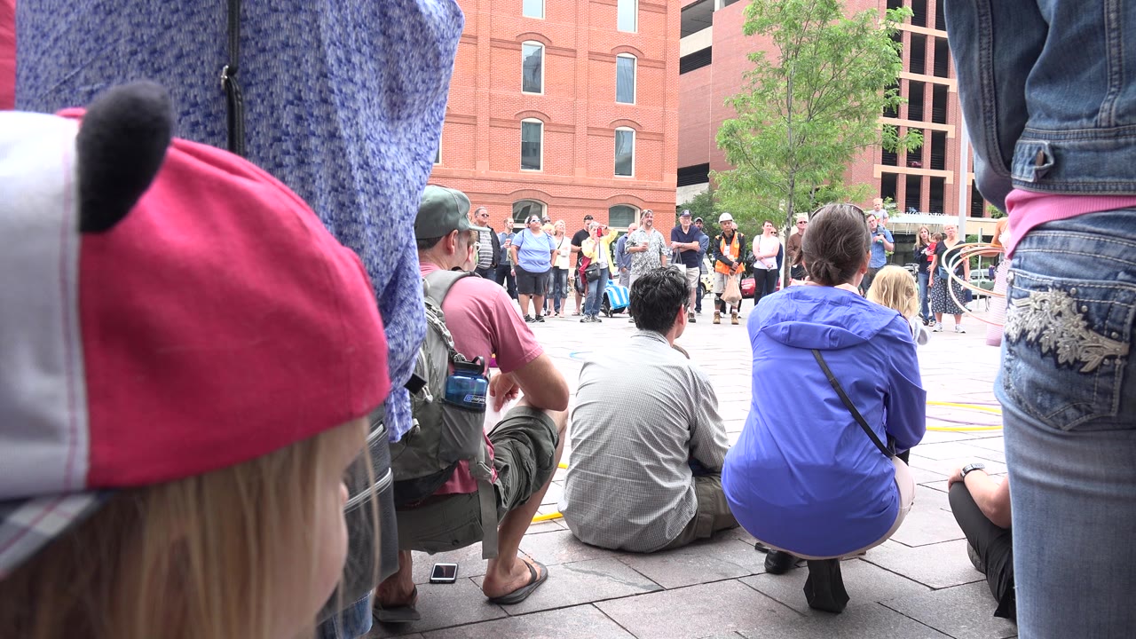 Grand Junction, Colorado audience sits and stands around an open plaza while the hula hoop performer appears beyond the foreground spectators near red brick buildings.