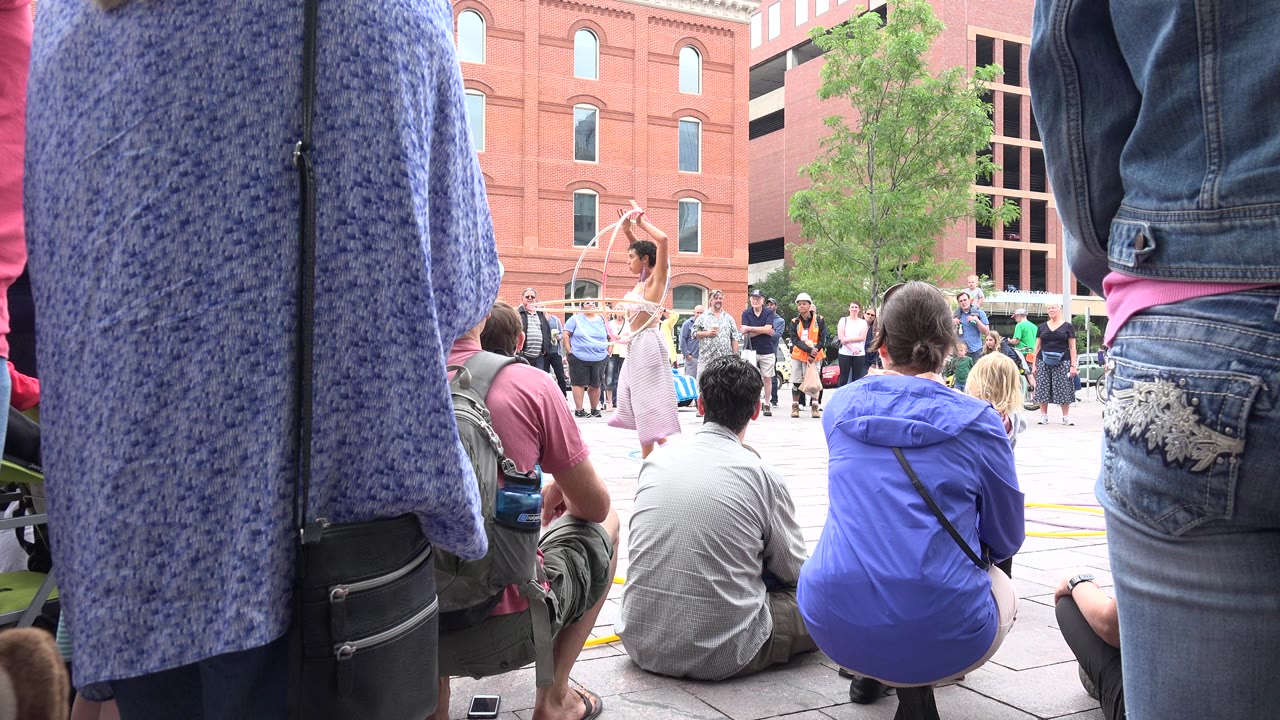 A woman in a light outfit performs with hoops while people sit and stand around the edge of a Grand Junction plaza.