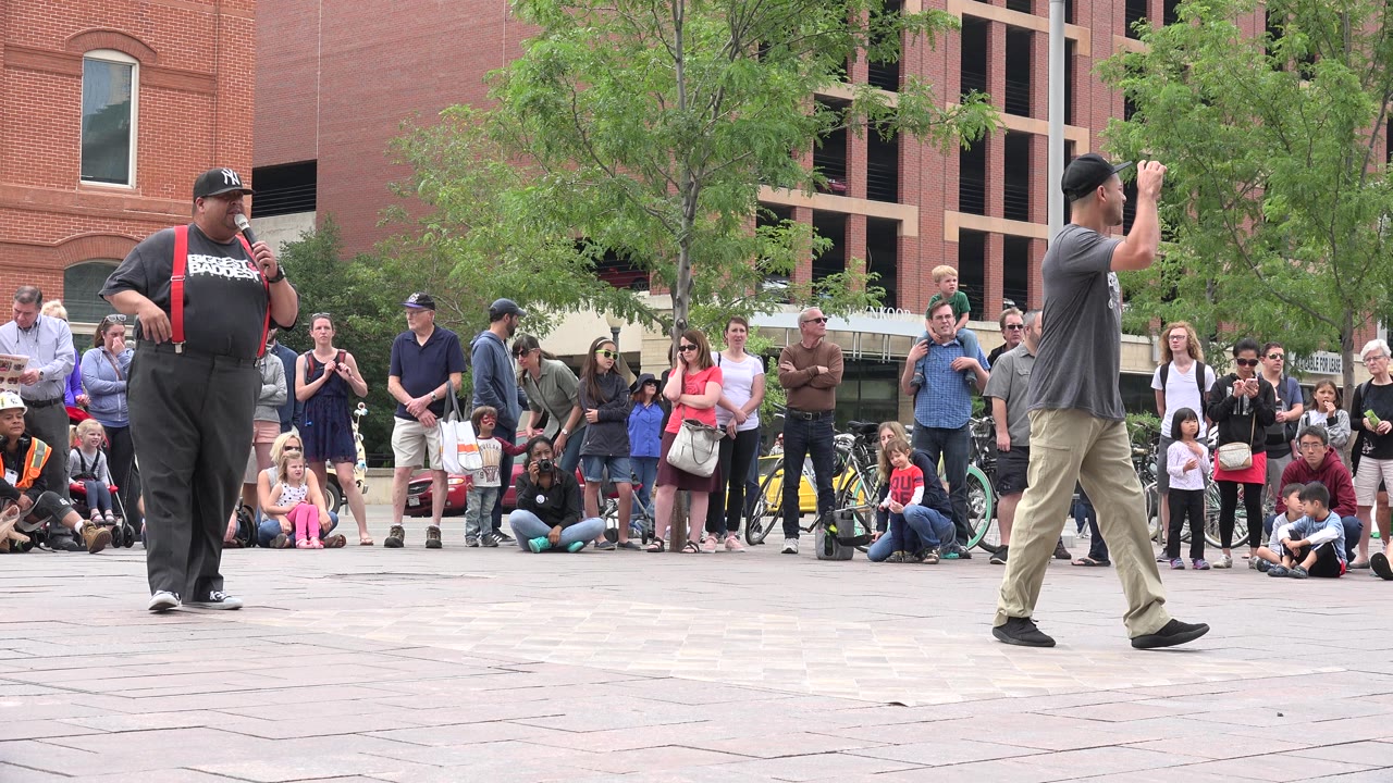In Grand Junction, Colorado, a performer in black with red suspenders stands at left while another man faces a crowd gathered around the plaza.