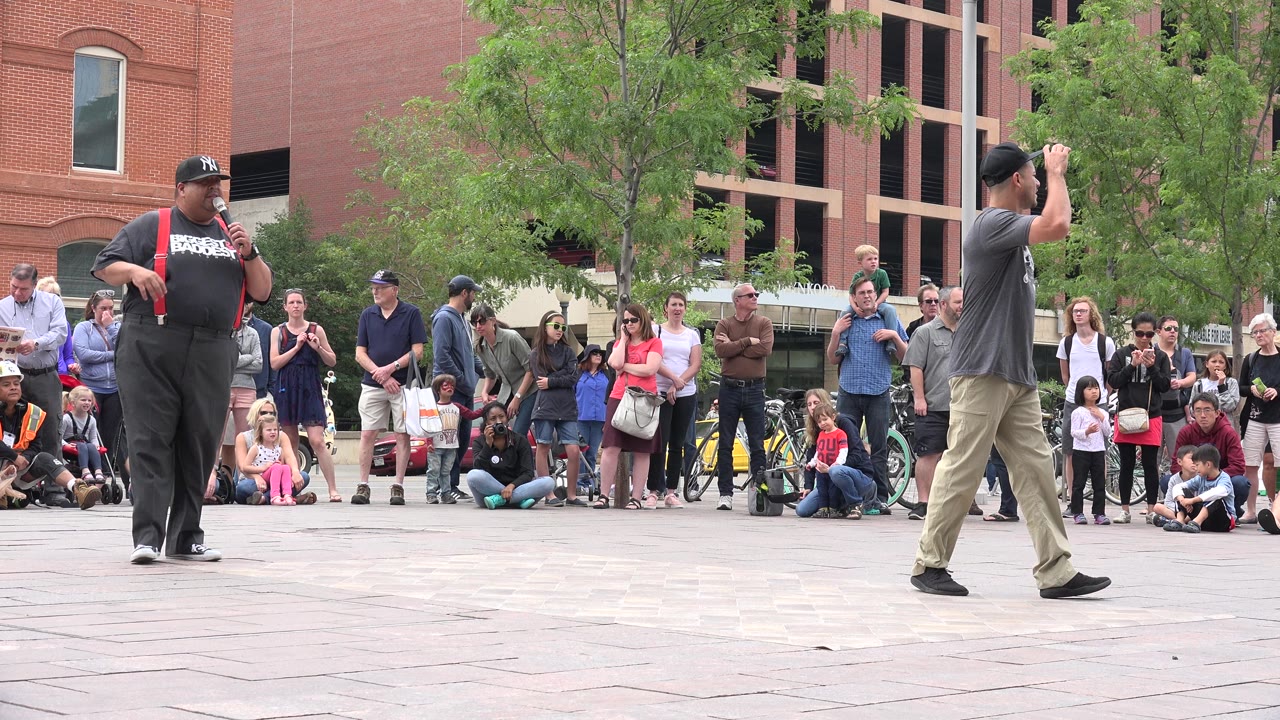A man in a gray shirt and cap crosses the open plaza foreground while spectators, children, trees, and brick buildings fill the background in Grand Junction, Colorado.