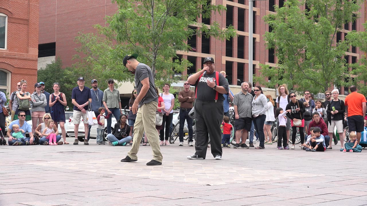 Two male performers stand in front of a gathered Grand Junction audience, with one looking down and the other holding a microphone near the crowd line.