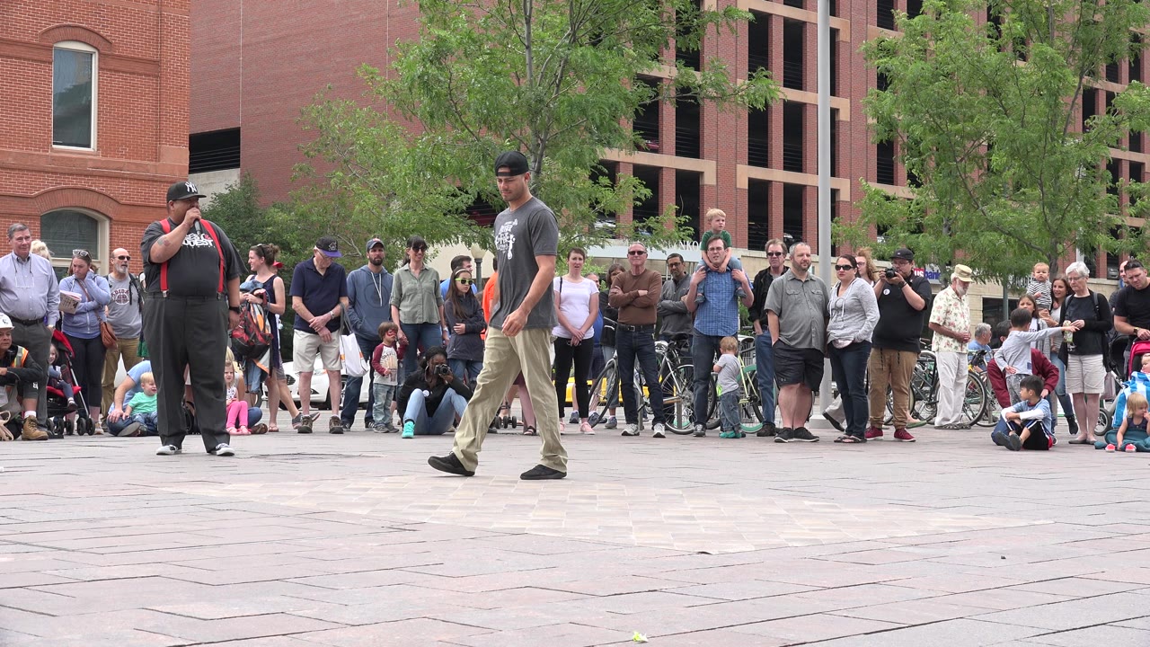A performer in a gray shirt walks across the open plaza as the microphone-holding performer stands near the left side and the Grand Junction crowd watches behind them.