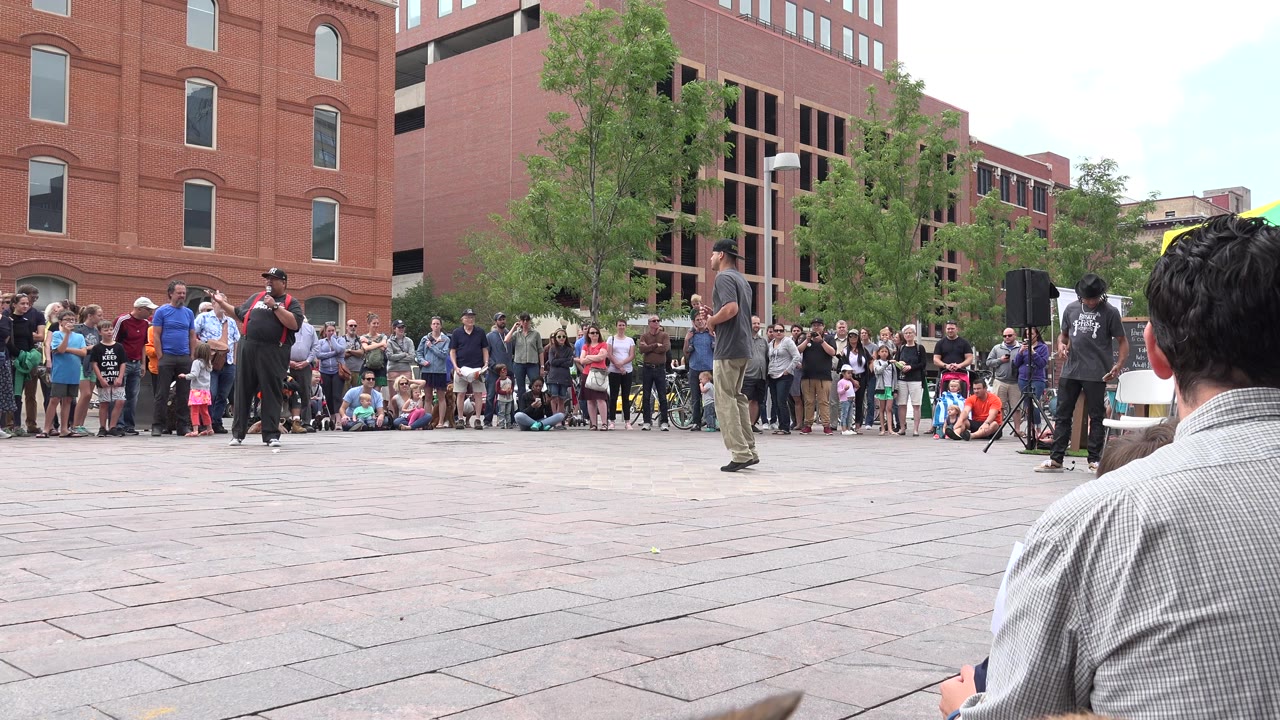 A Grand Junction, Colorado street performer stands in an open plaza with spectators, children, trees, speakers, and red brick buildings behind him.