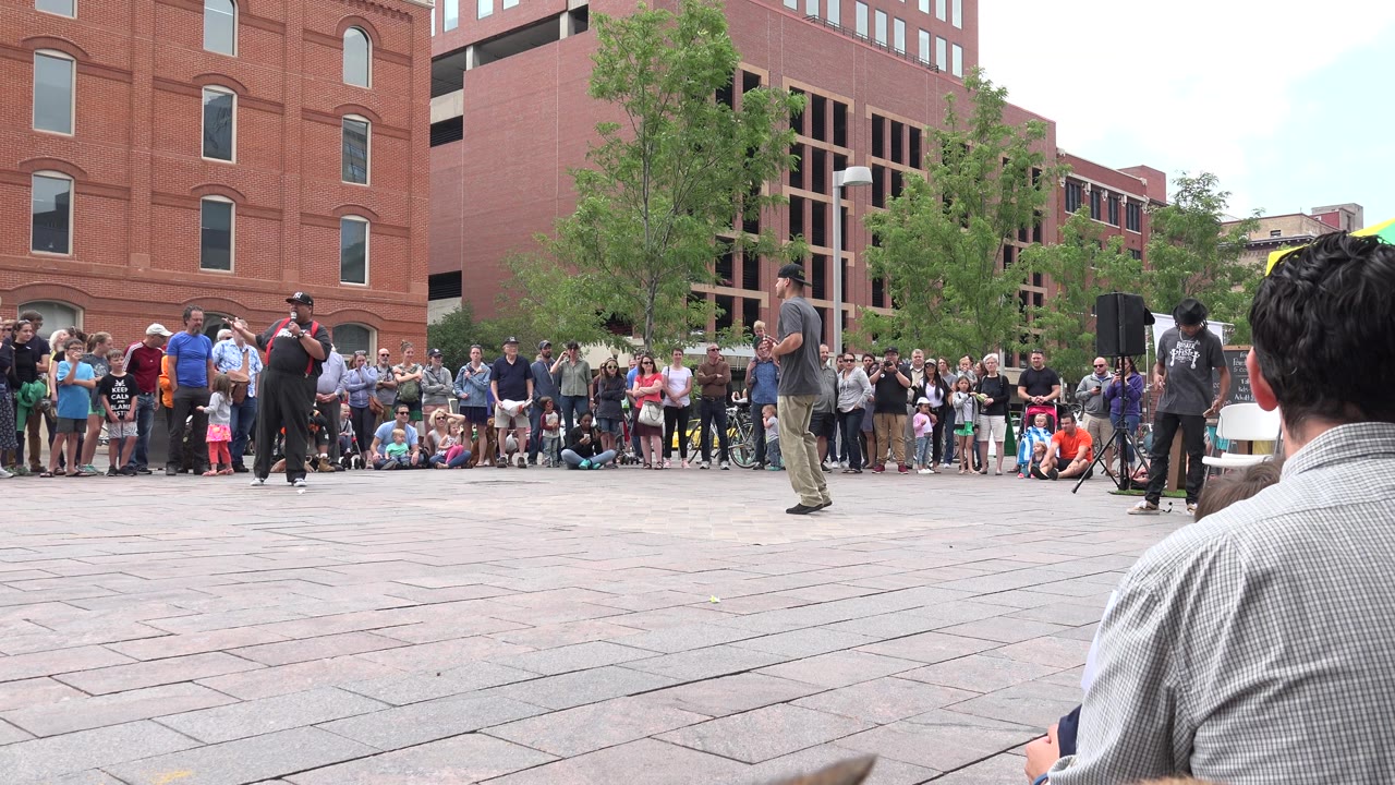 The performer walks across the Grand Junction plaza while a line of spectators watches from the edge of the paved open space.