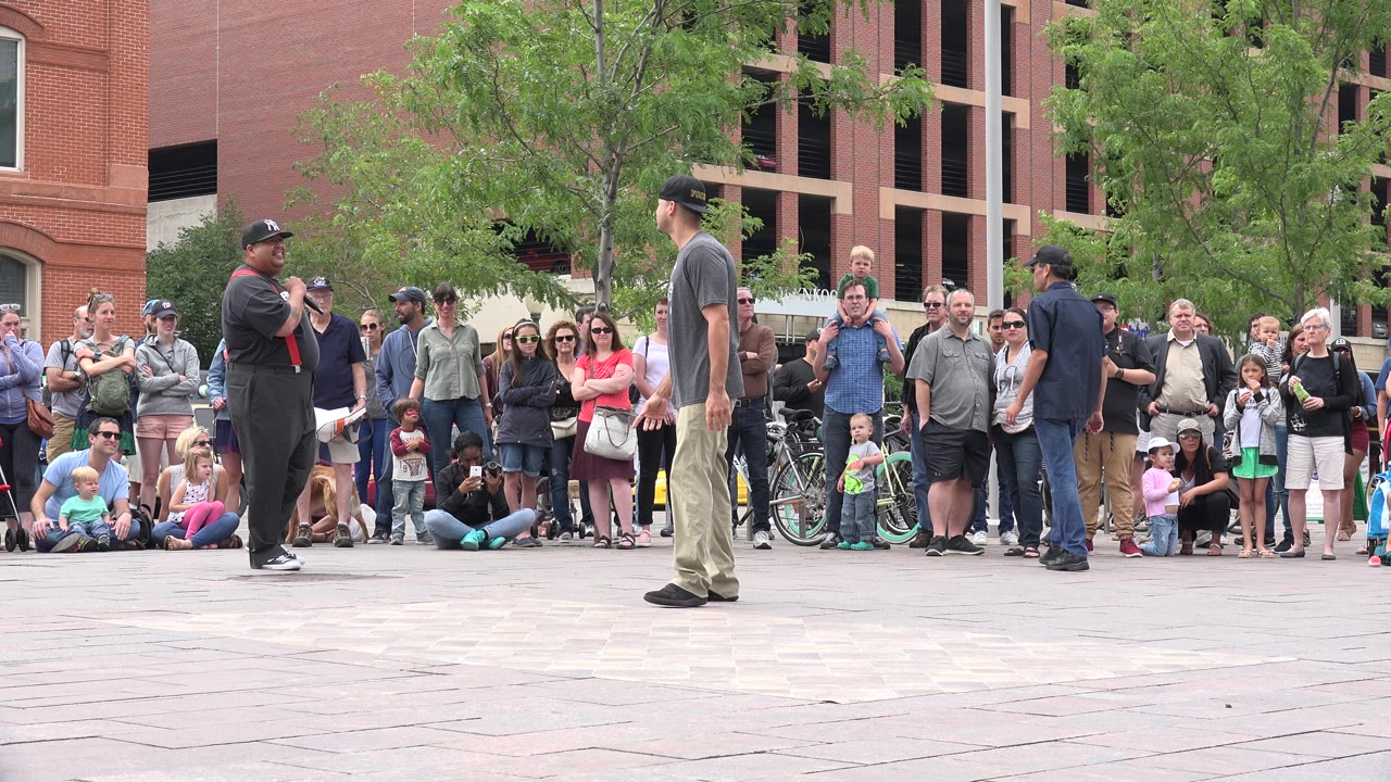 A dancer in a gray shirt stands near a microphone-holding performer as spectators line the background in the Grand Junction, Colorado plaza.