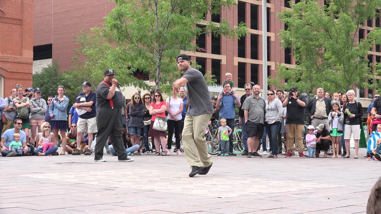 The dancer lands into a step near another performer in dark clothing while a standing crowd watches in the Grand Junction plaza.