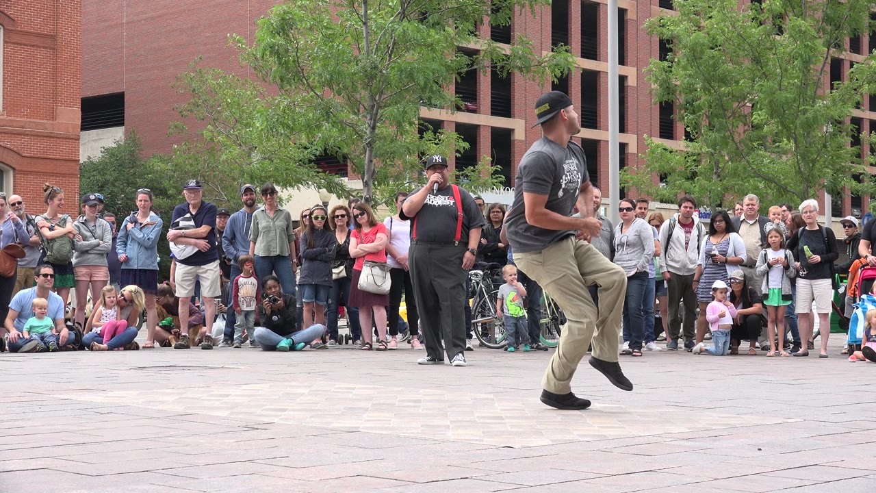 The dancer turns his body to the side while spectators, children, trees, and brick buildings remain visible behind him in Grand Junction, Colorado.