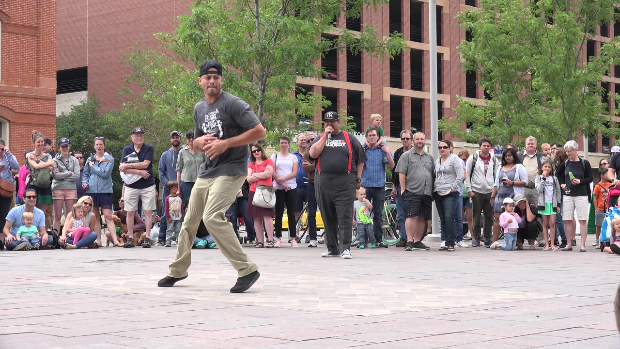 The dancer faces forward in mid-step on the sidewalk, with a dense line of spectators behind him near a red brick building and leafy tree in Grand Junction, Colorado.