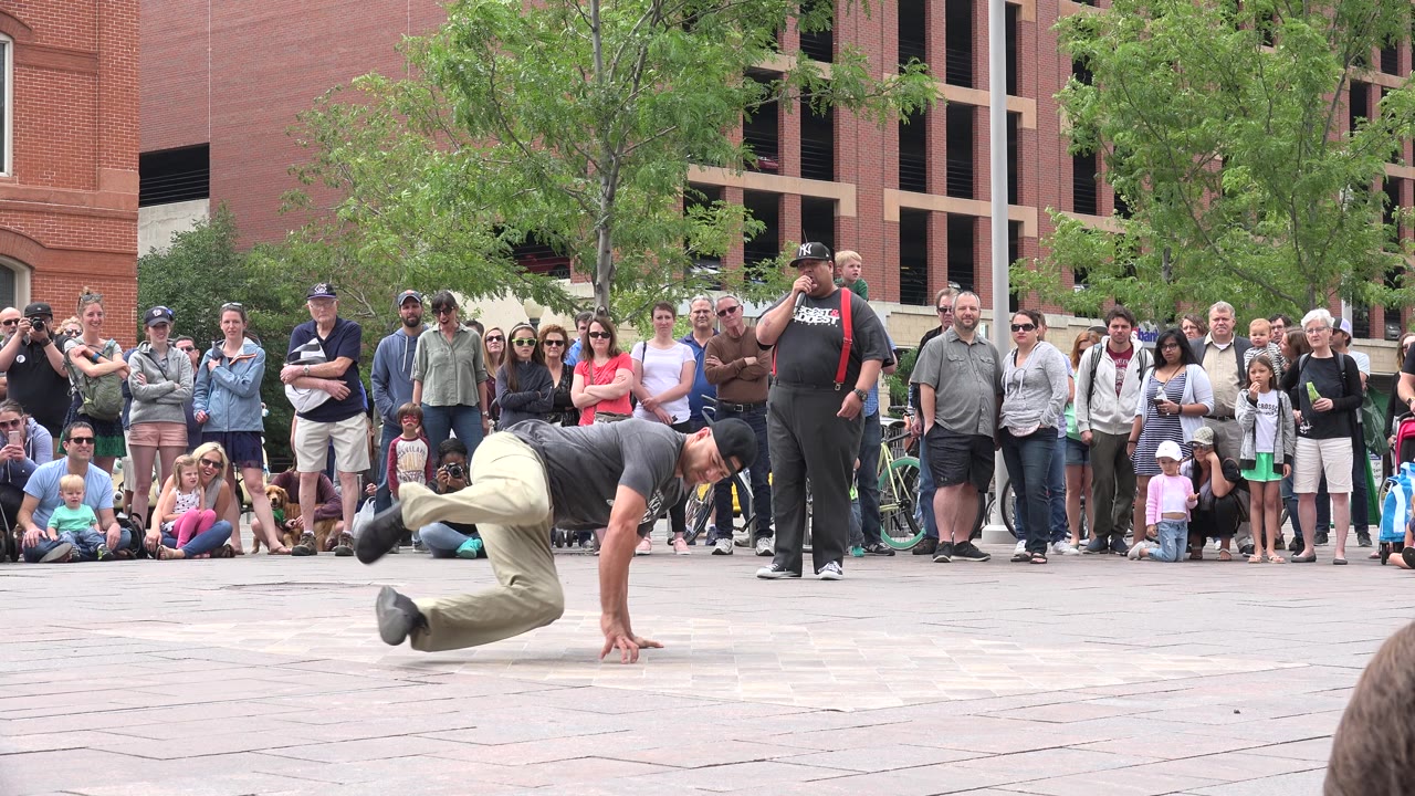 In Grand Junction, Colorado, the dancer lifts both legs off the ground in a breakdance move as a crowd stands and sits behind him near trees and a brick building.