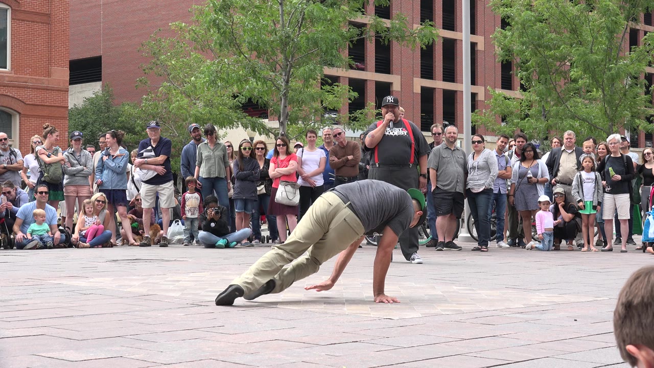 A break dancer in a gray shirt balances low on one hand on a paved plaza in Grand Junction, Colorado, with spectators lined up behind.