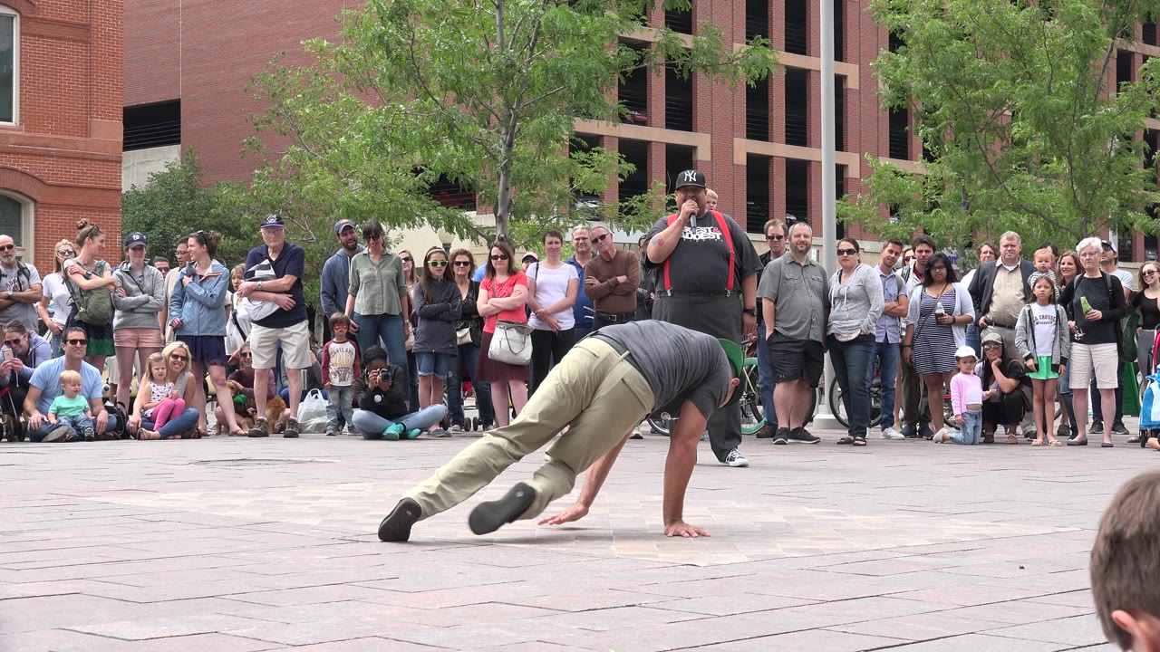 The dancer extends both legs close to the plaza surface while supporting their body with one arm, with the Grand Junction crowd still facing the performance.