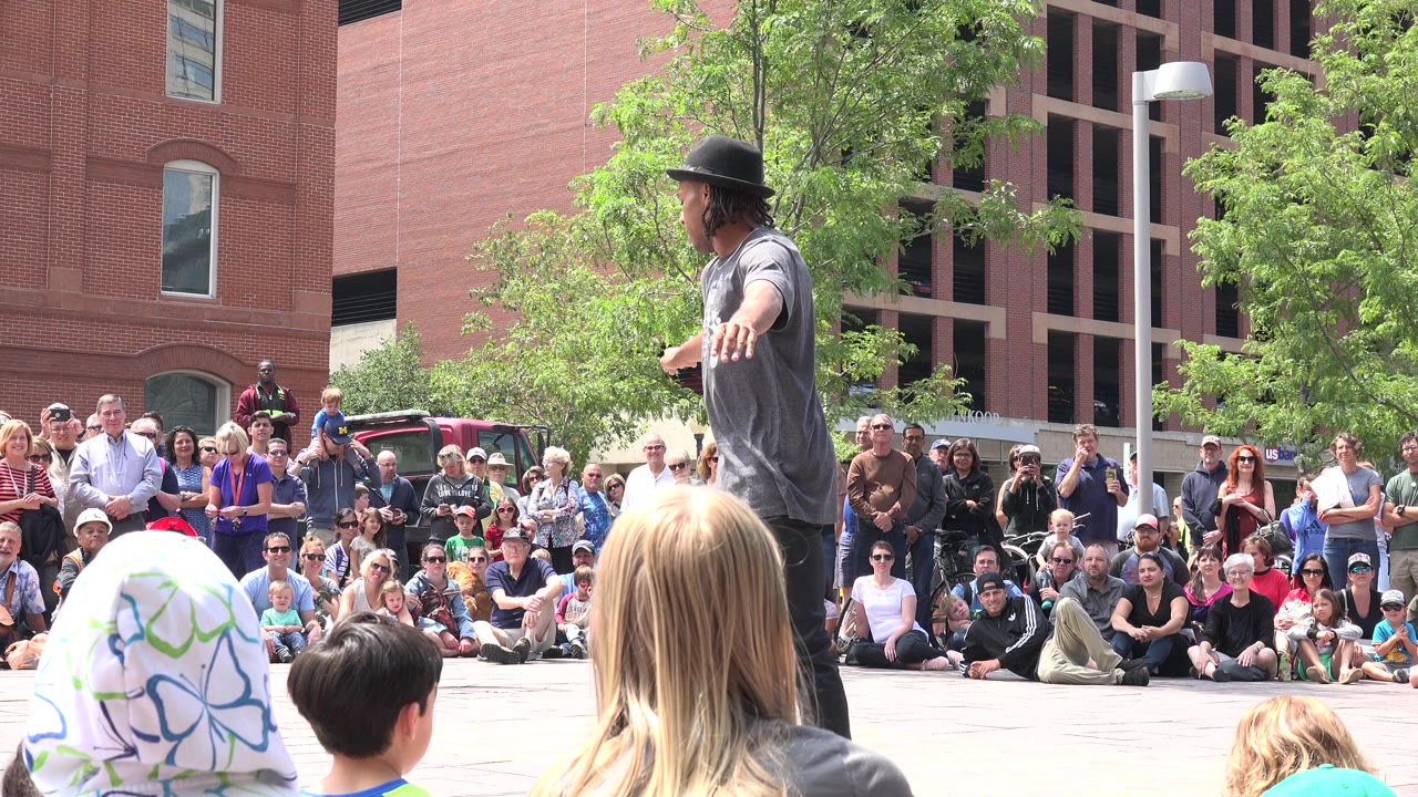 The performer keeps one arm out toward the crowd as spectators sit and stand across the Grand Junction plaza under bright daylight.