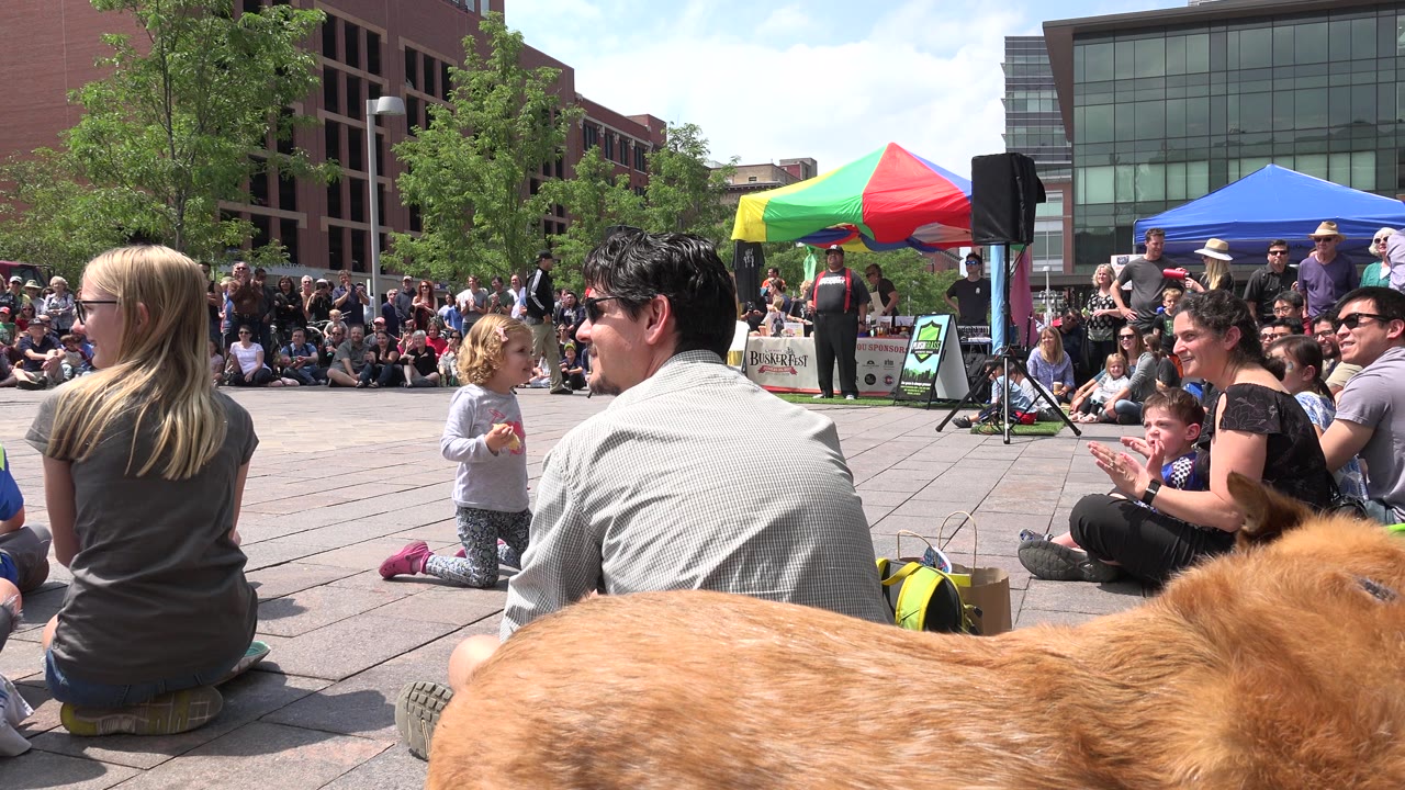 Seated crowd watches a plaza performance with tents and brick buildings behind them.