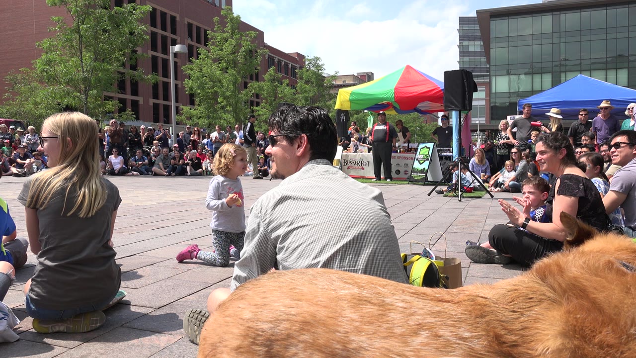 A tan dog fills the lower foreground while adults and children sit on the plaza in Grand Junction, Colorado, watching the same performance area near colorful tents and brick buildings.
