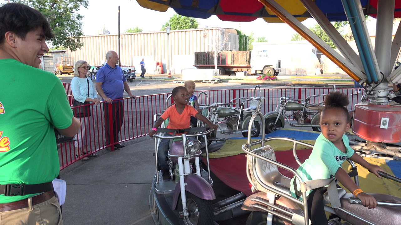 At 0:00 in Grand Junction, Colorado, two children sit in motorcycle-style merry-go-round cars while a green-shirted ride operator stands at the left edge and adults watch behind red railings.