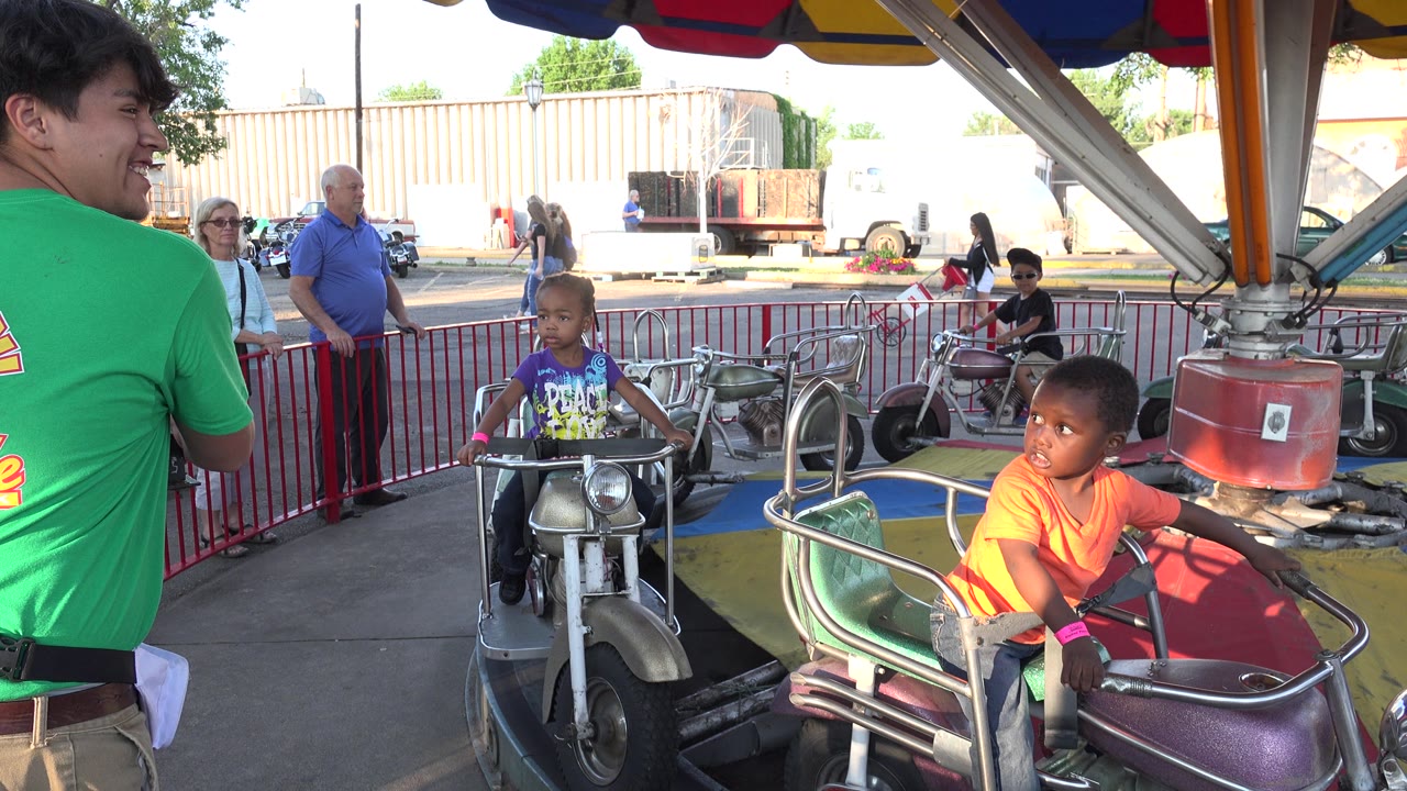 Children ride motorcycle-shaped amusement cars while adults stand behind a red barrier.