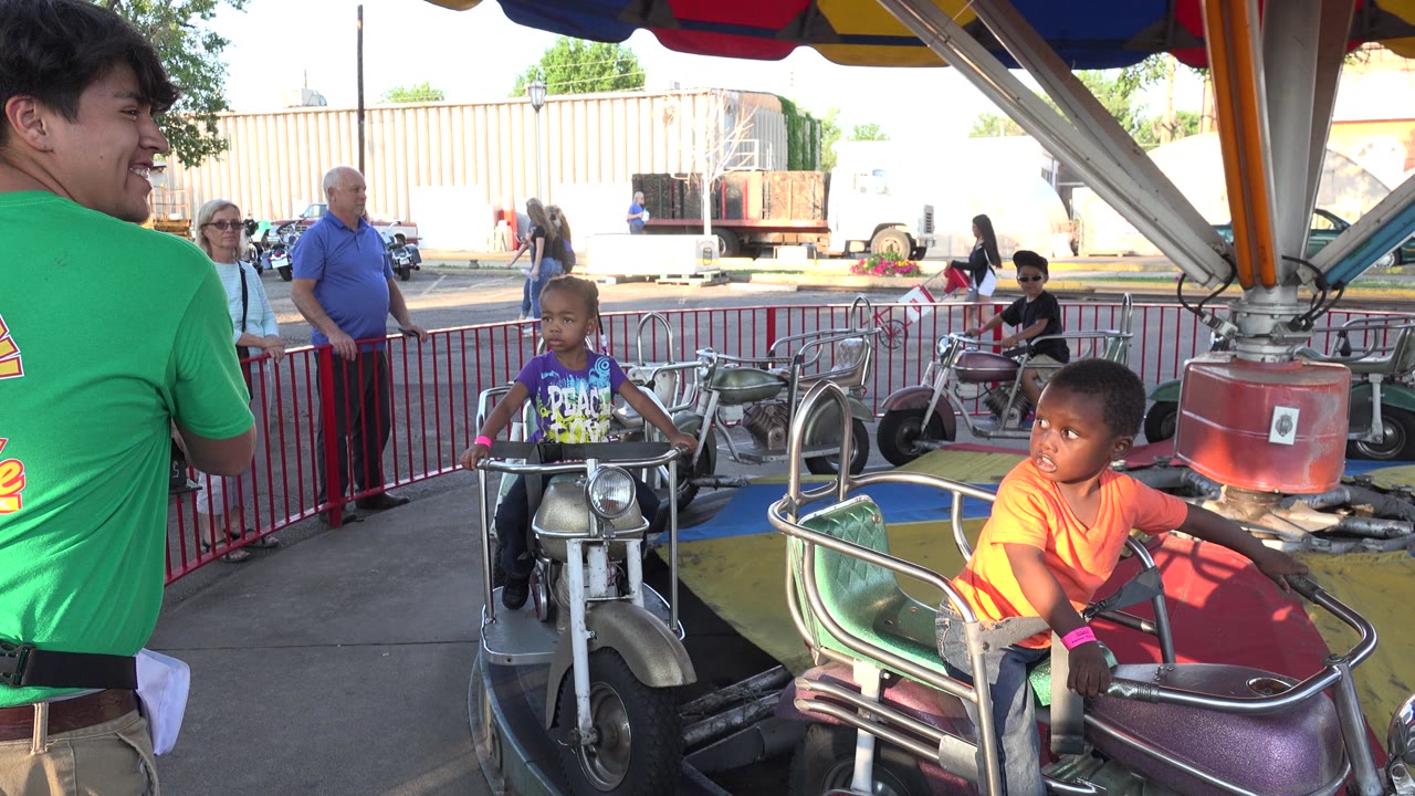 At 0:01 in Grand Junction, Colorado, a boy in an orange shirt rides closest to the camera while another child on a motorcycle car approaches from the center under the striped canopy.