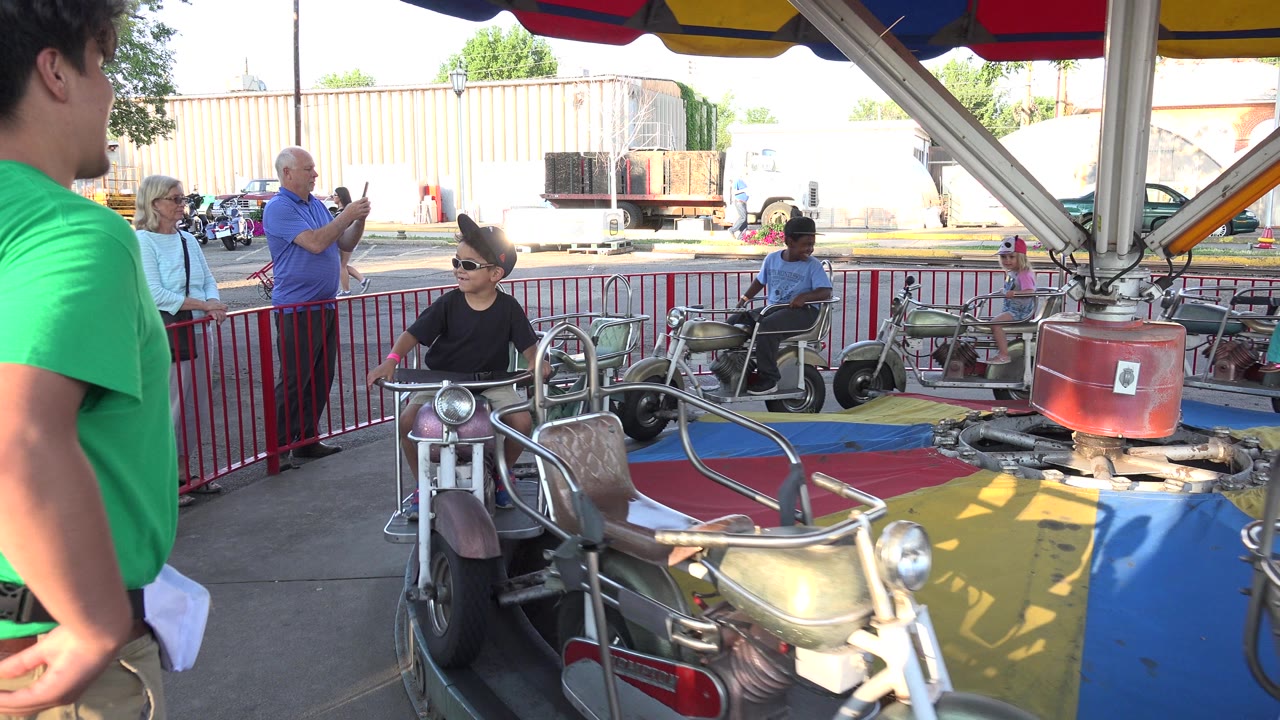Child seated on a bicycle-style fairground ride near adult spectators.