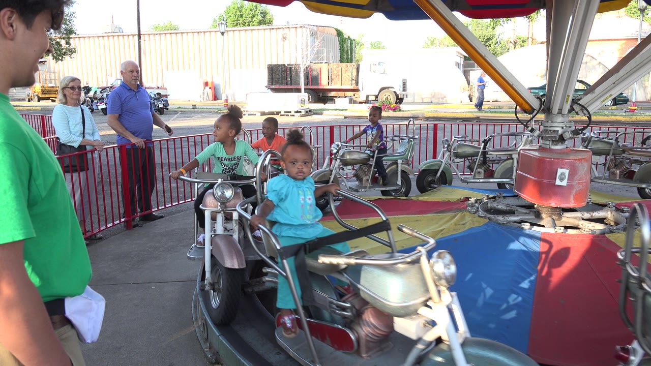 Children on motorcycle-style amusement ride with adults behind a red fence.
