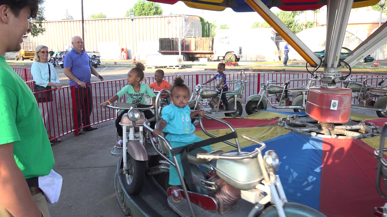 A child in a turquoise shirt rides near the foreground as other children circle behind on the Grand Junction amusement ride, with adults watching beyond the railing.