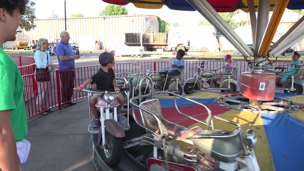 Children ride small carousel motorcycles while adults stand behind a red rail.