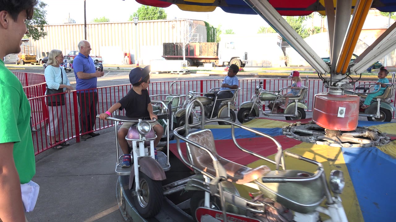 One second into the Grand Junction, Colorado ride clip, a child in a dark shirt passes close to the camera on a maroon motorcycle-style carousel car.