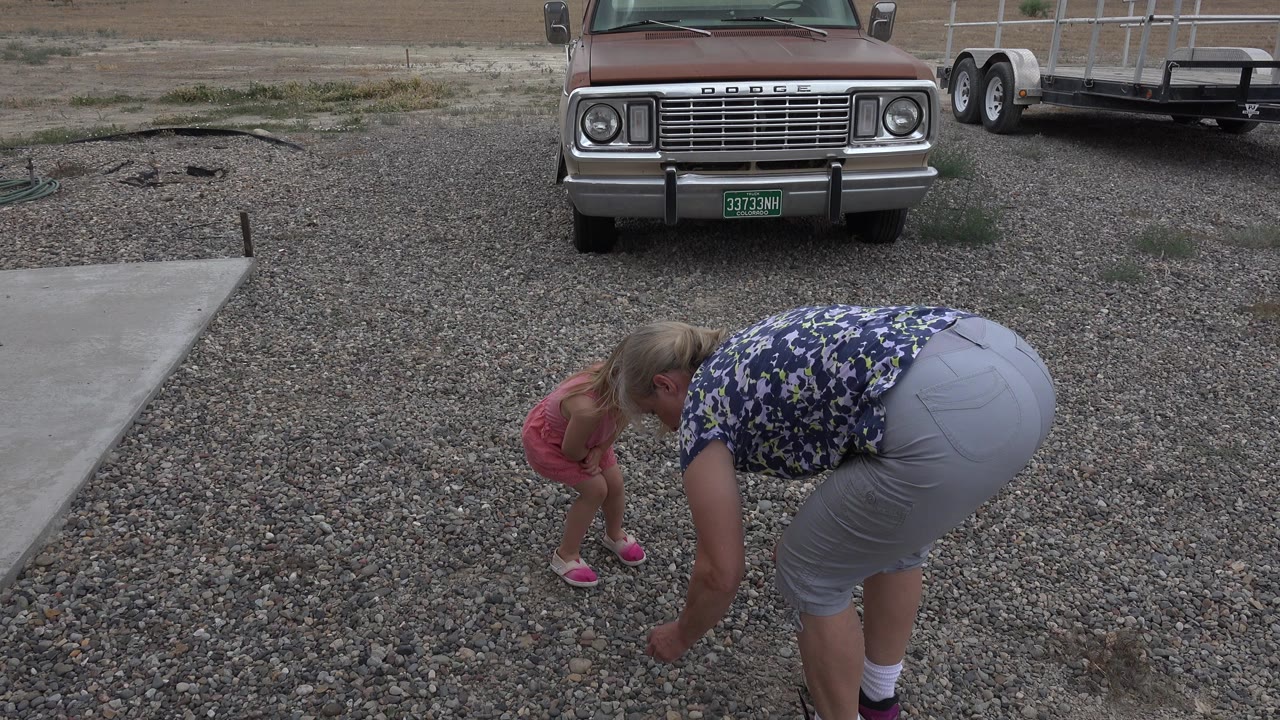 In Grand Junction, Colorado, a woman bends near a young girl in a pink dress on a gravel driveway, with a Dodge truck and utility trailer in the background.