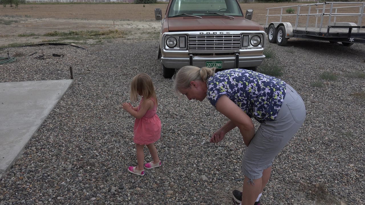 Woman bending near a young girl on a gravel driveway beside a parked truck.