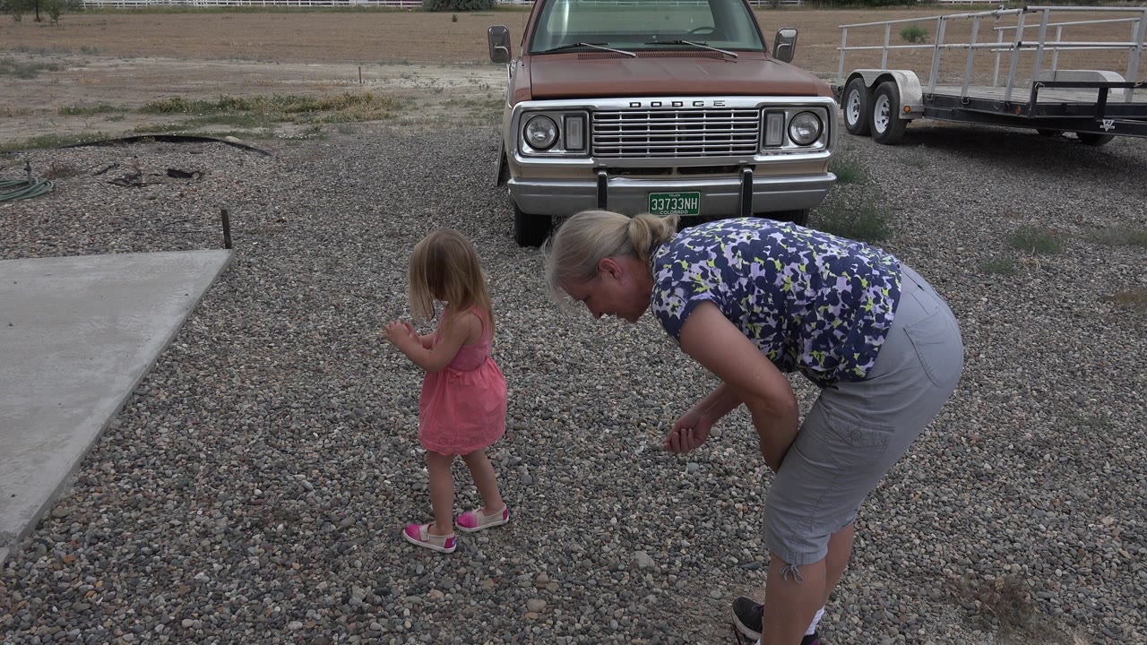 The Grand Junction driveway scene continues as the woman stays bent over and the girl stands turned slightly away, with gravel, the concrete pad, pickup truck, trailer, and dry field visible.