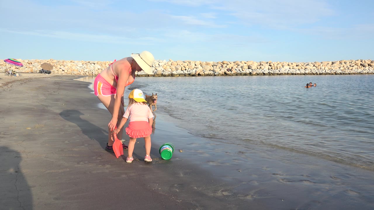 At 0:01, the adult remains bent toward the child as the green bucket lies near the waterline, with the rock breakwater and small distant swimmers visible across the background.