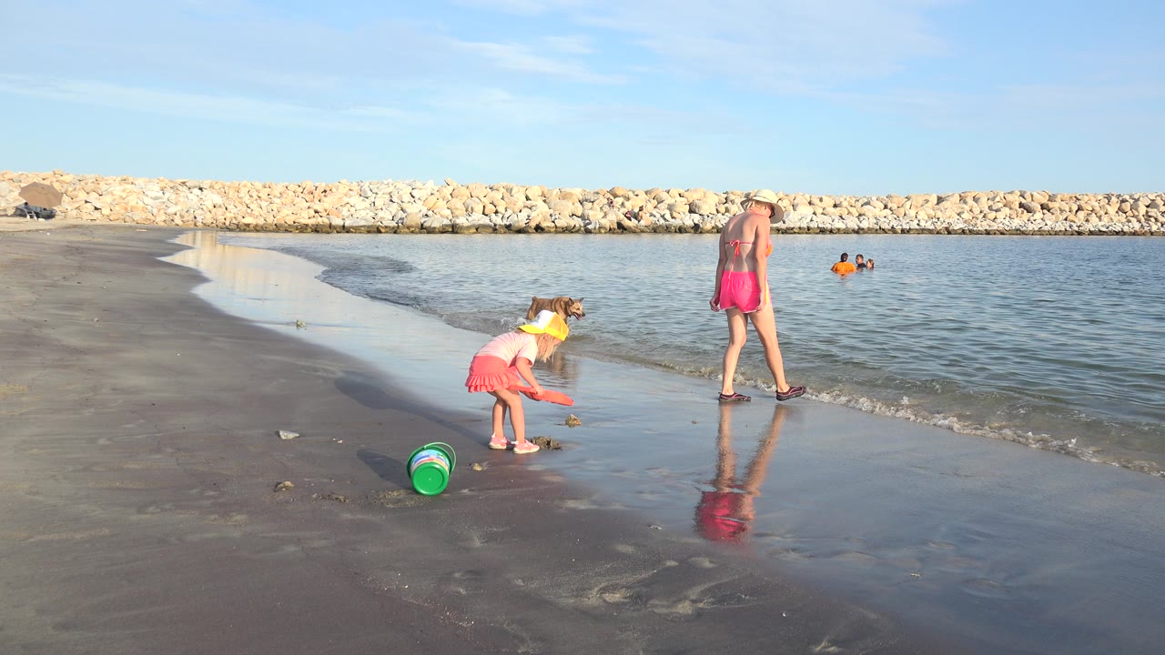 Child plays on wet sand near a green bucket while an adult stands at the waterline.