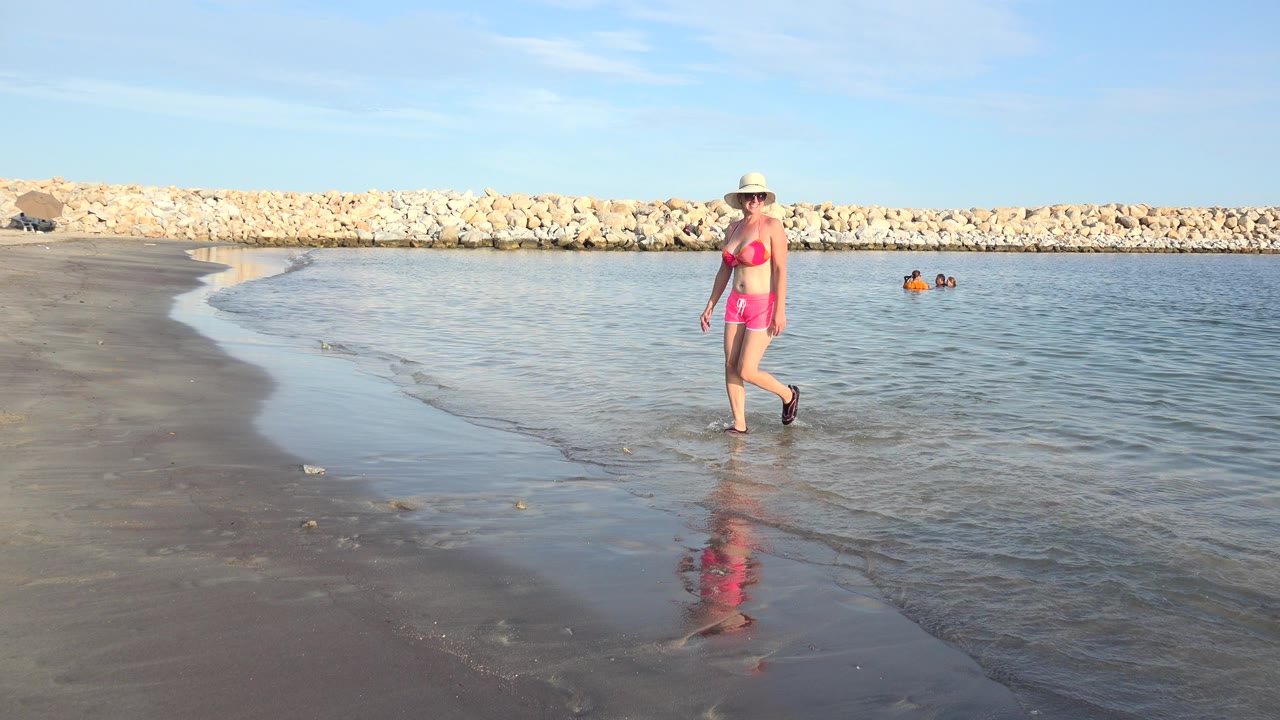 Woman in pink swimwear walks along wet beach beside calm water.