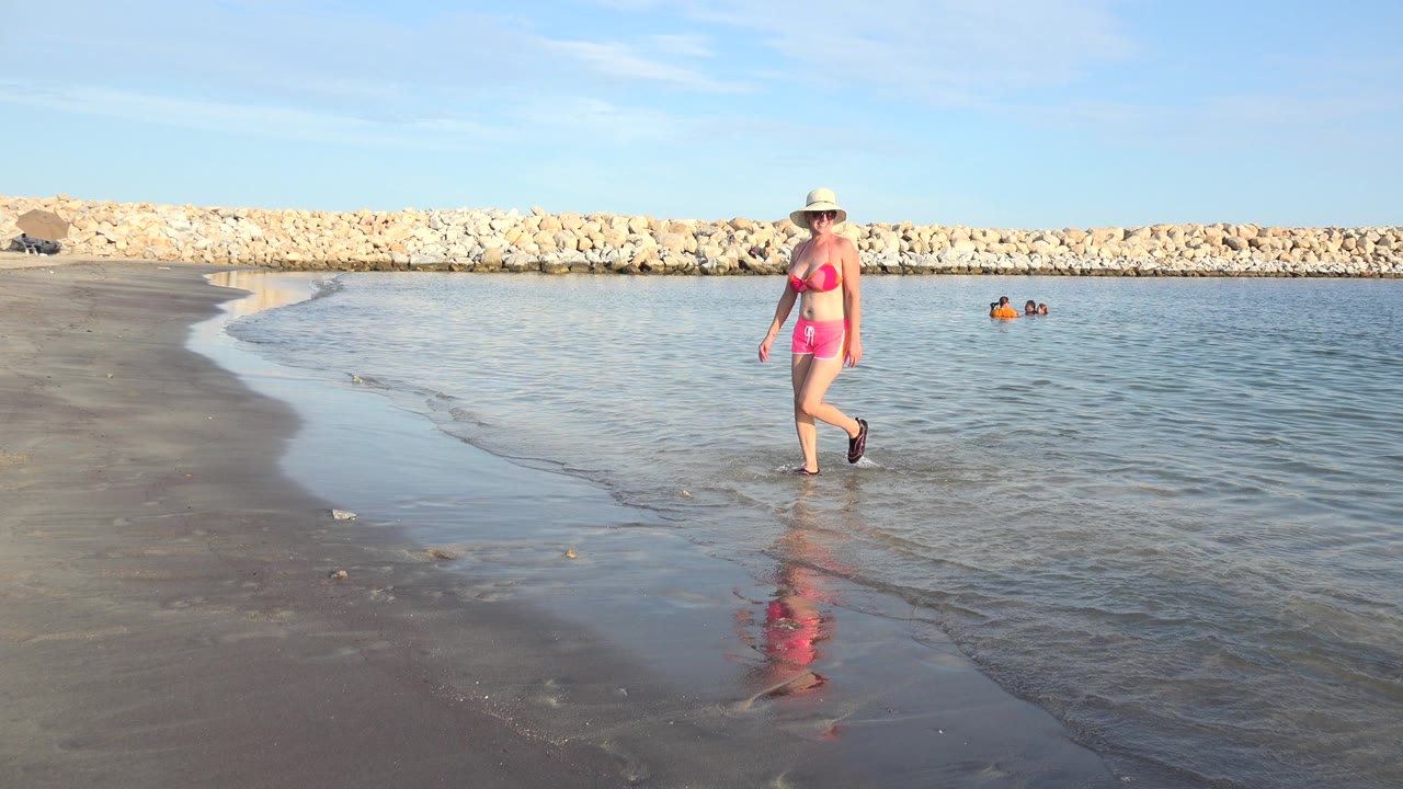 The woman remains centered on the Grand Junction shoreline, walking over reflective wet sand while small waves meet the beach and distant swimmers sit in the water.