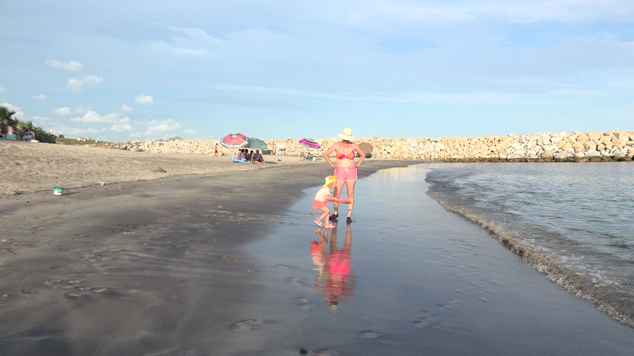 In Grand Junction, Colorado, a woman in a sun hat stands near a child crouching at the wet shoreline, with umbrellas, a rock barrier, and bright water in the background.
