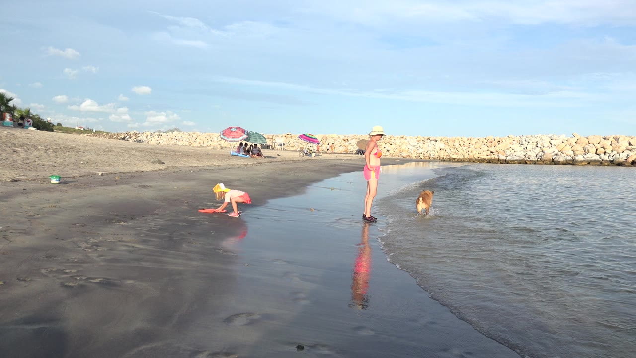At 0:00 in Grand Junction, Colorado, a child in a yellow hat bends over wet sand while a woman stands by the water and a small dog faces the ripples near a low rock barrier.