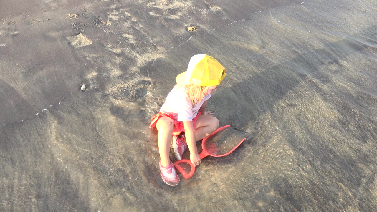 Blonde child in a yellow cap crouching on wet sand with an orange shovel.