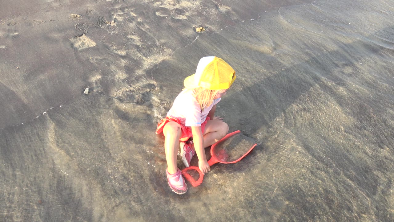 The child remains crouched in Grand Junction, Colorado, with the yellow cap catching bright light and the orange shovel resting on rippled wet sand.