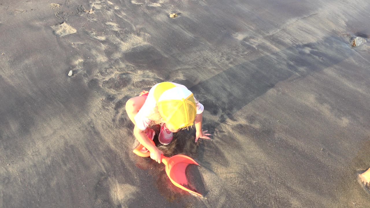 Child in a yellow cap kneeling on wet sand with an orange shovel.