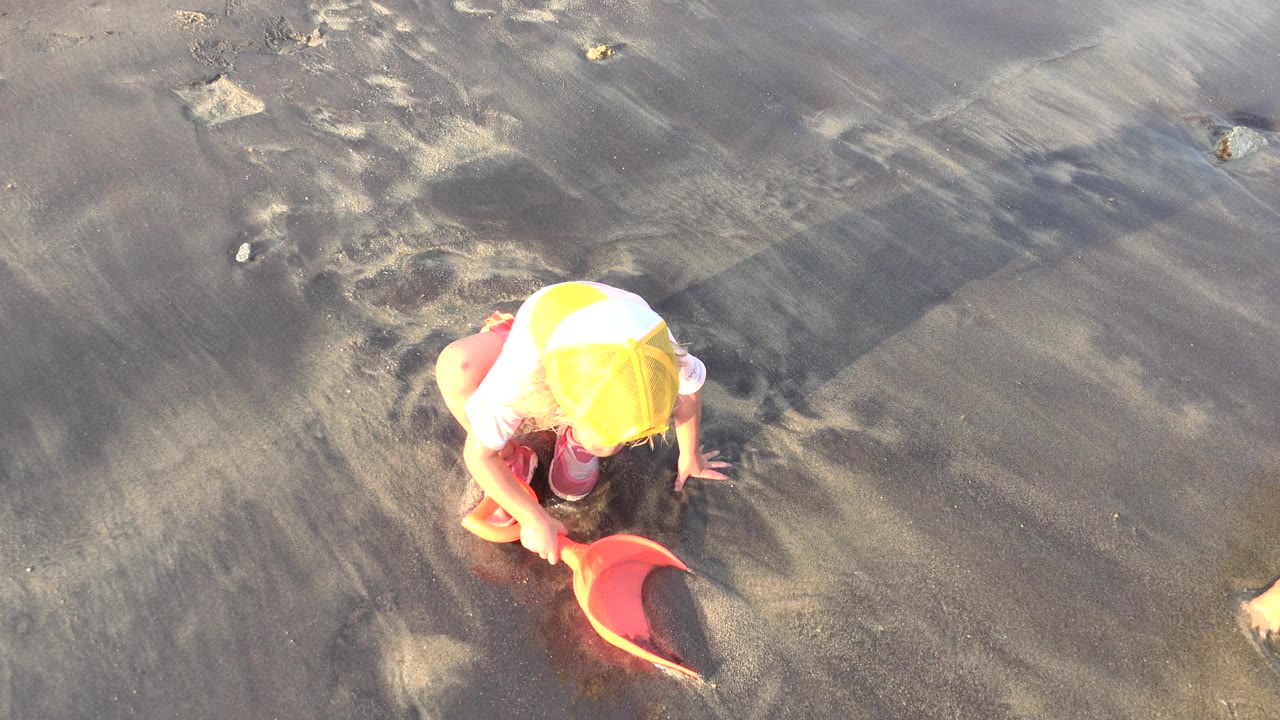 At 0:01 in Grand Junction, Colorado, the child stays crouched with the orange shovel angled across wet sand as long shadows stretch behind them.