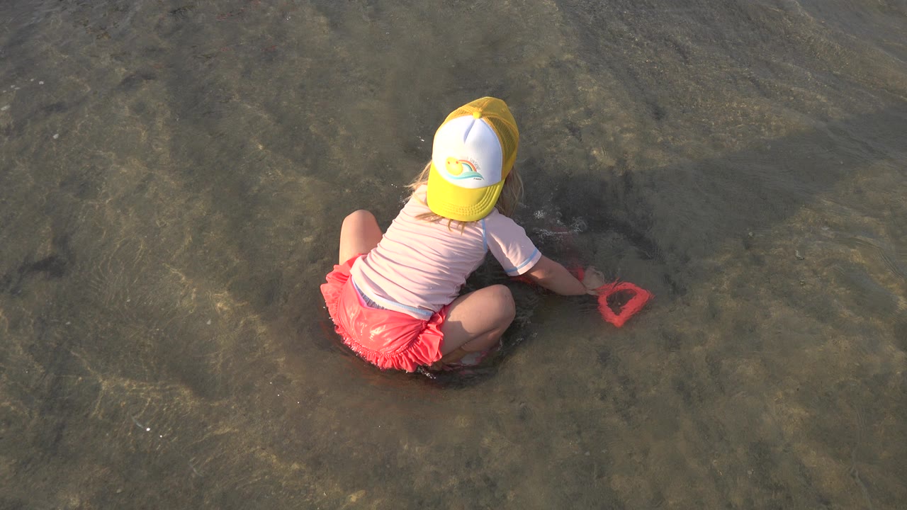 Young girl crouching in shallow water with a red shovel.