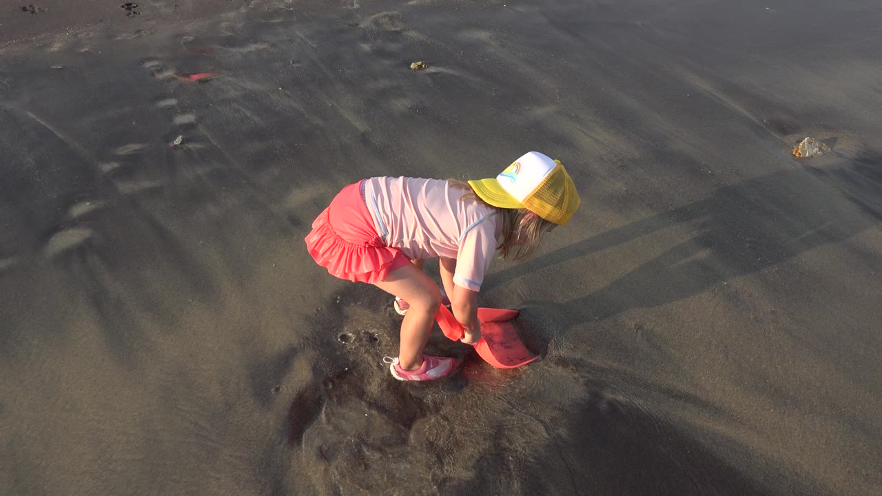 Girl in a yellow cap bends over wet sand with a pink scoop.