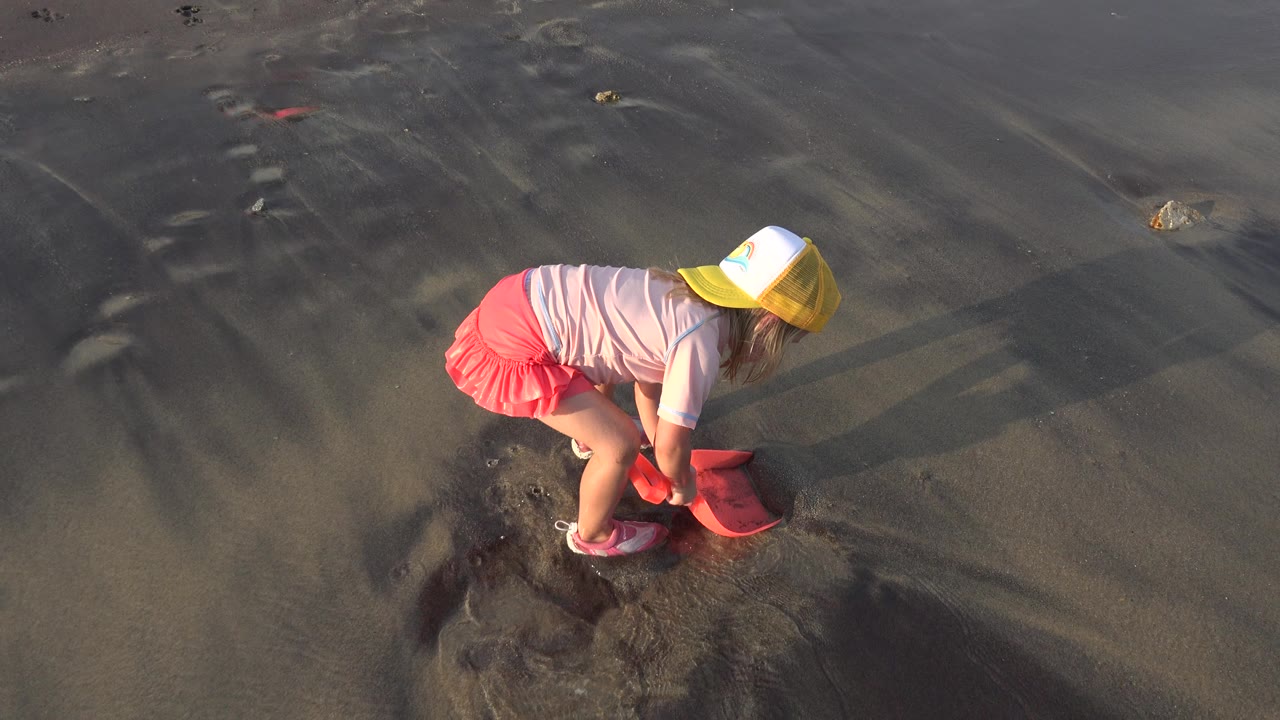 At 0:01, the child stays bent over the wet shoreline sand, with her yellow cap catching the light and the pink scoop pressed close to the ground.